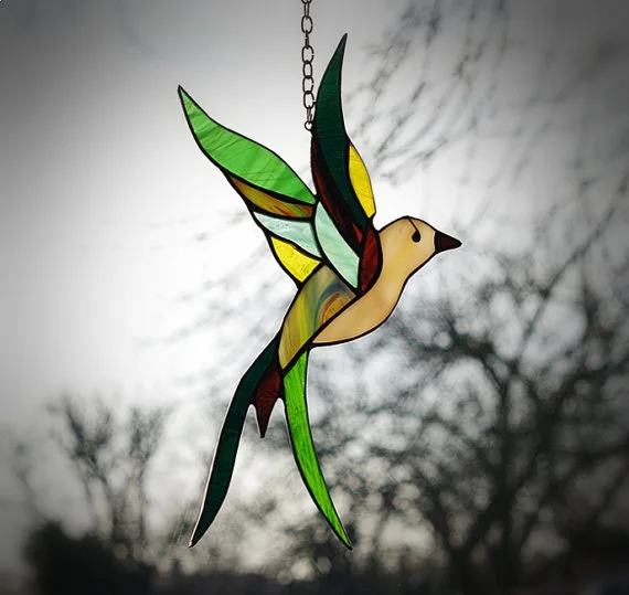 Un petit colibri en verre coloré, photographié en suspension devant une fenêtre.