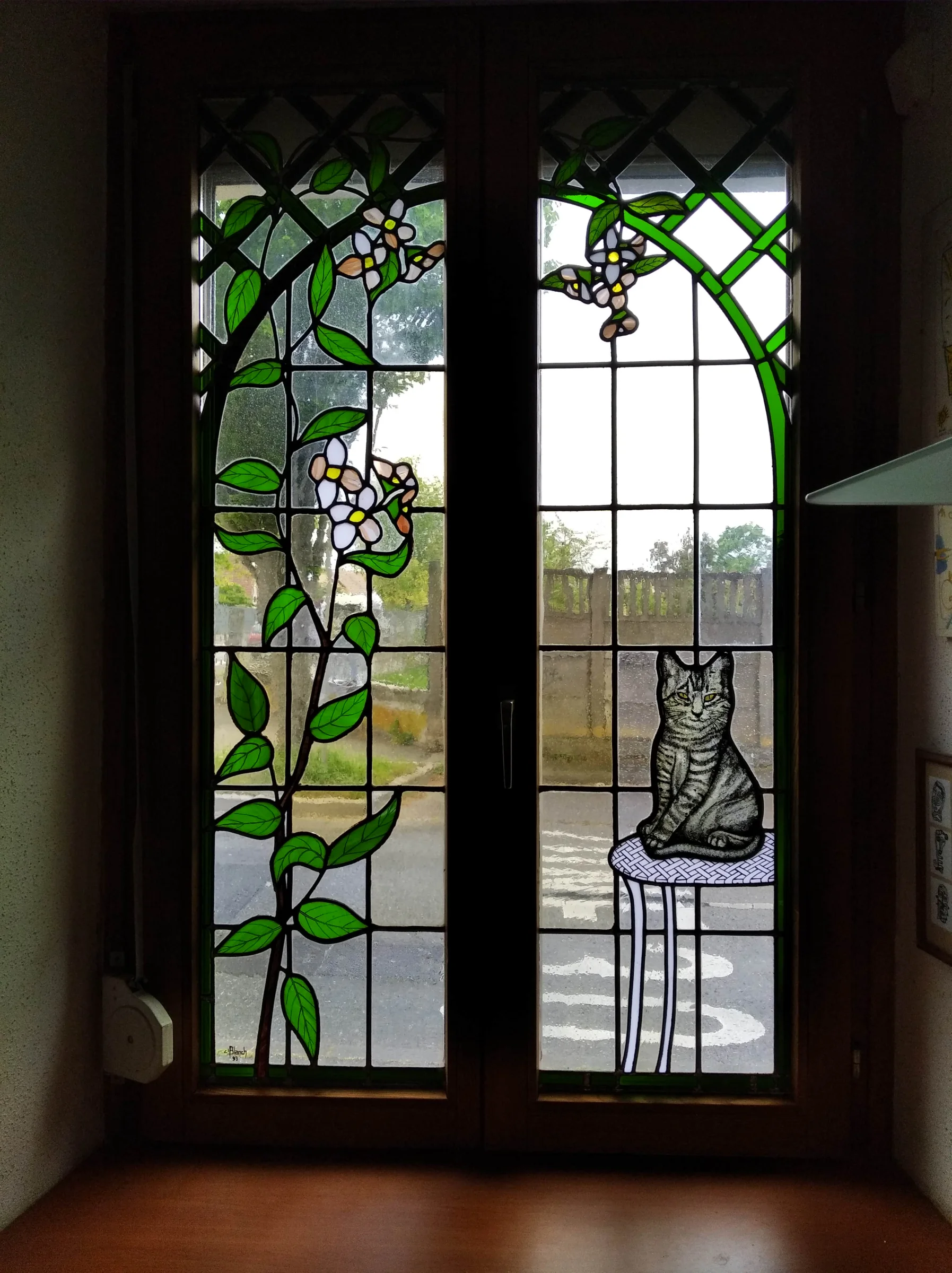 Une fenêtre à deux panneaux associant un chat, des fleurs et un feuillage vert.