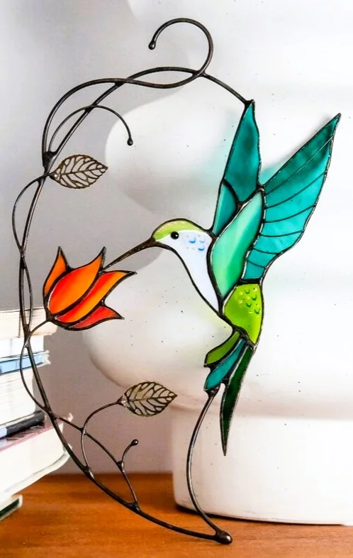 Un colibri vert près d’une fleur orange, dans une composition en verre présentée sur un support.